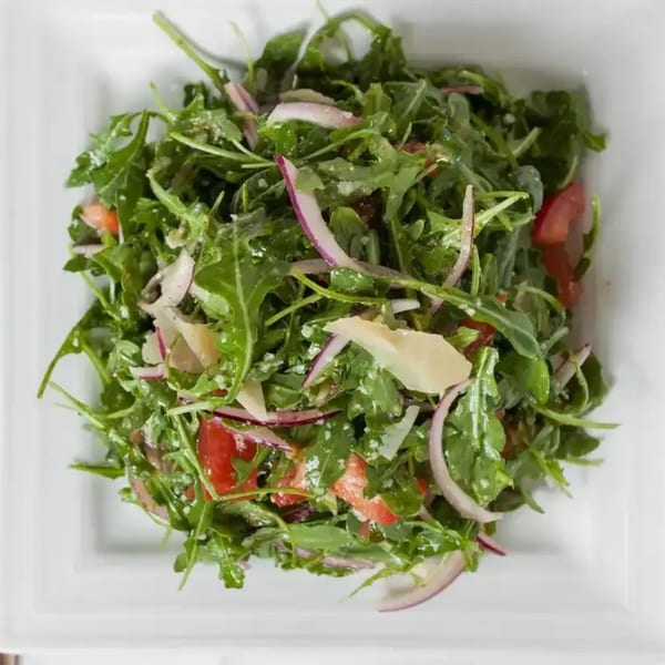 Arugula salad with red onions, tomatoes, and parmesan cheese in a white square bowl.