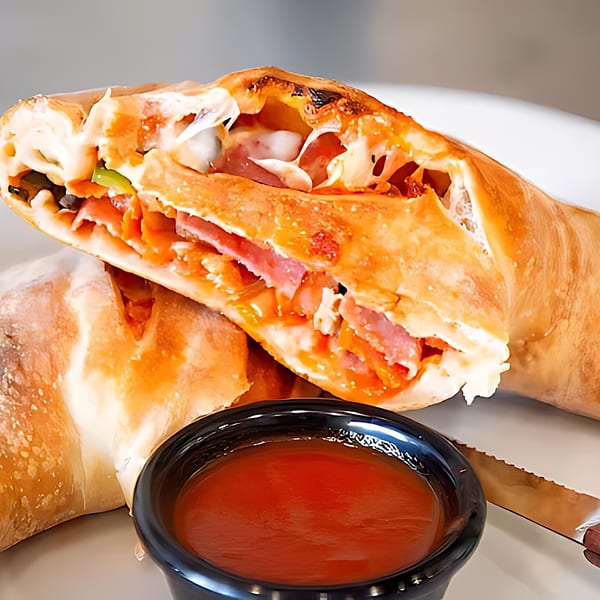 A golden-brown calzone, sliced open to reveal melted cheese, ham, and vegetables, with a side of marinara sauce.